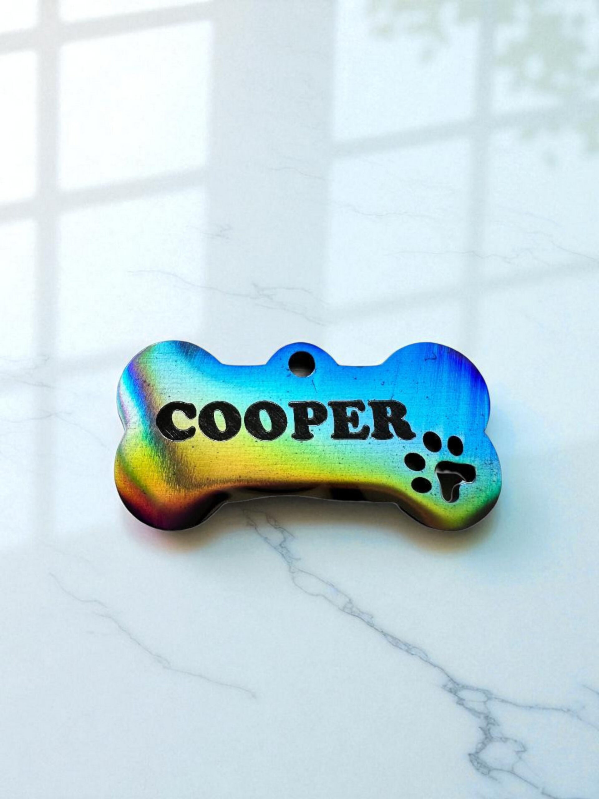 A colorful pet ID tag in the shape of a bone, engraved with the name 'COOPER' and a paw print design, placed on a light marble surface.