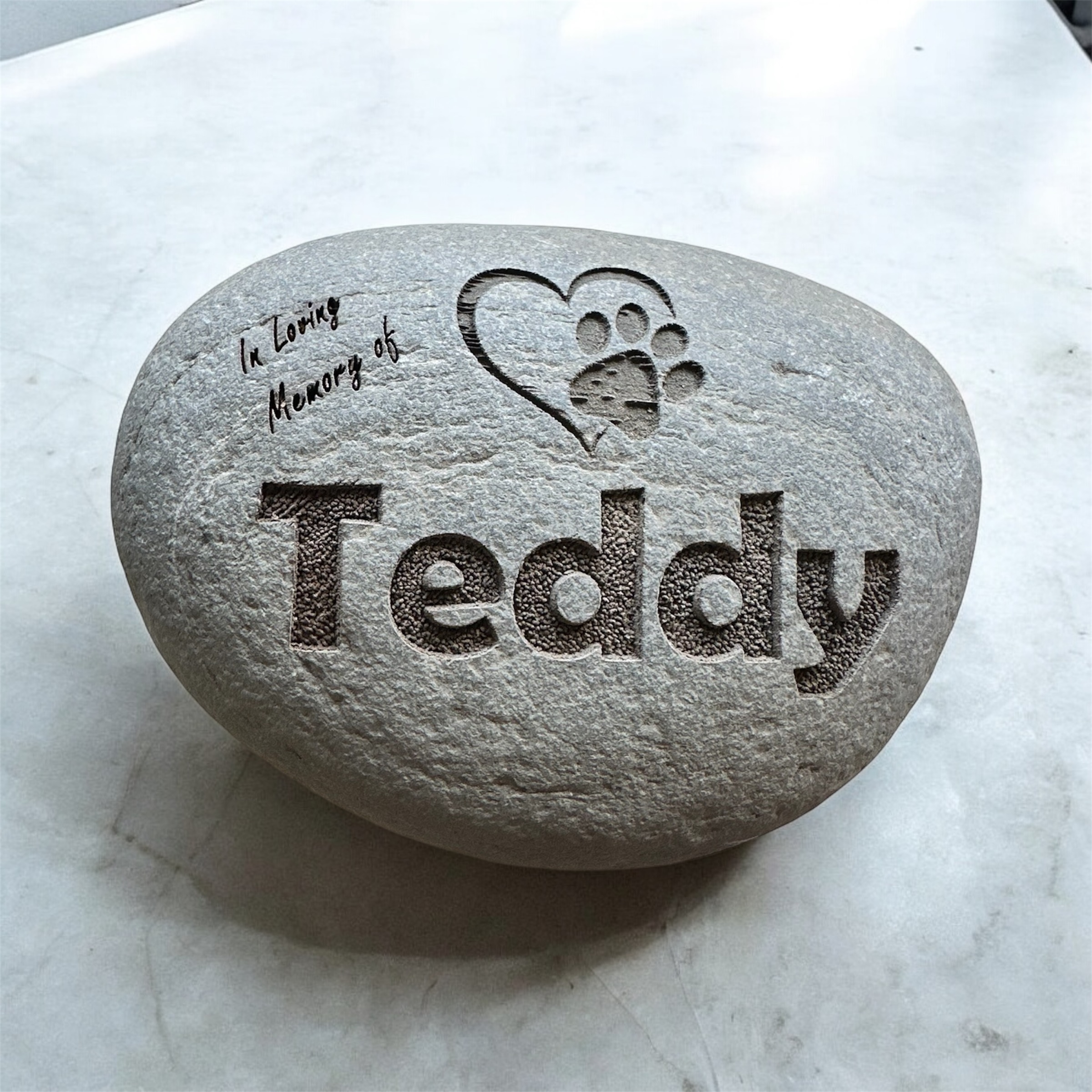 A memorial stone engraved with the name 'Teddy' and a heart design, with the phrase 'In Loving Memory of' at the top.
